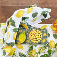 Table setting with lemon-patterned tablecloth, napkins, and glass dish on a wooden surface.